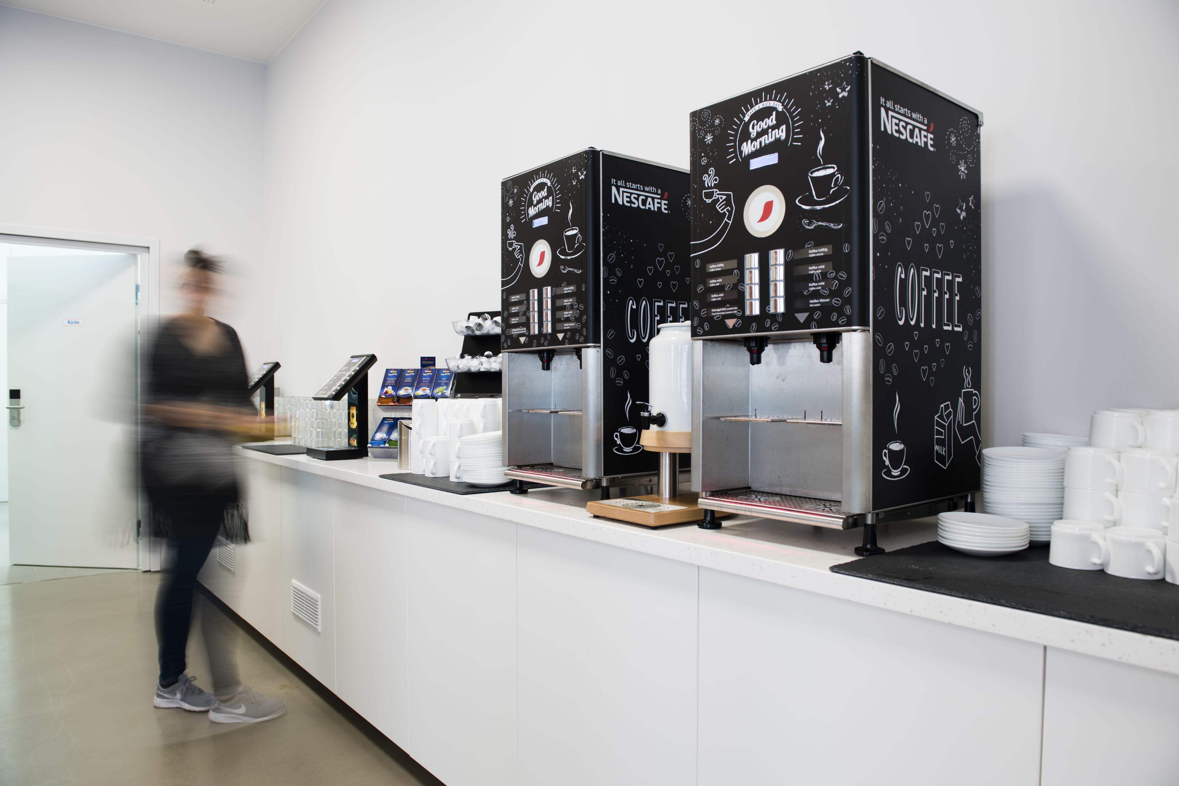 Un espace dédié au café, avec plusieurs machines Nescafé, un comptoir où sont empilées des assiettes et des tasses, et une personne qui passe.