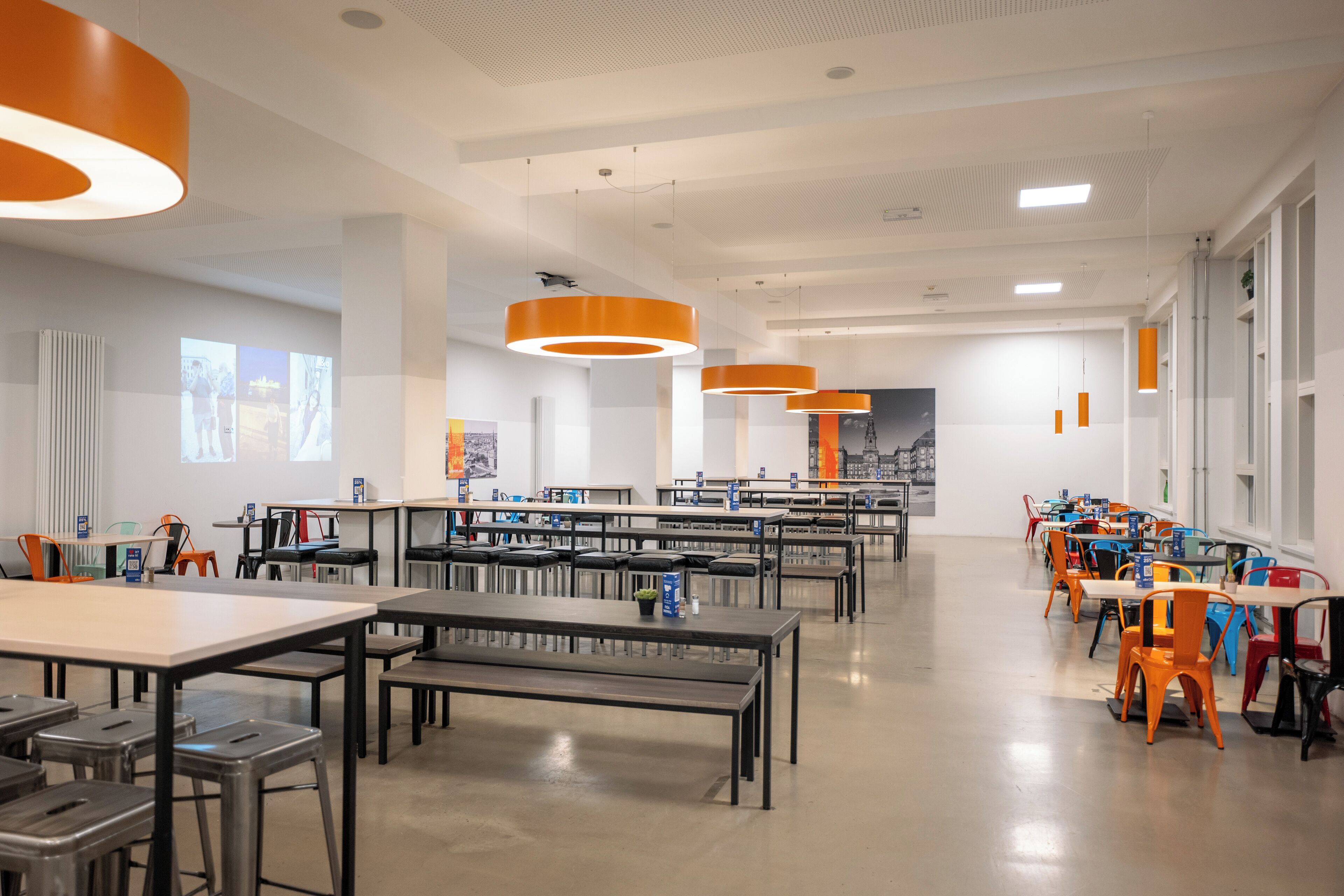 Une bibliothèque moderne dotée de nombreuses tables et chaises, de grandes fenêtres et de luminaires suspendus de couleur orange.