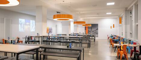 Une bibliothèque moderne dotée de nombreuses tables et chaises, de grandes fenêtres et de luminaires suspendus de couleur orange.