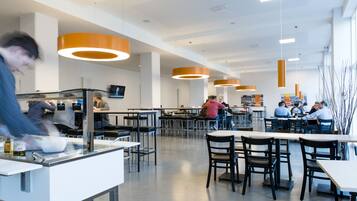 A modern cafeteria with high tables and chairs, overhead lighting, and a central serving area.