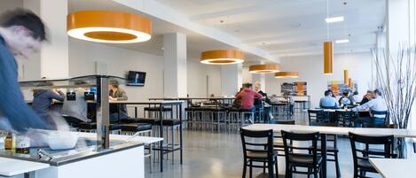 Une cafétéria moderne avec des tables et des chaises hautes, un éclairage suspendu et un espace de service central.