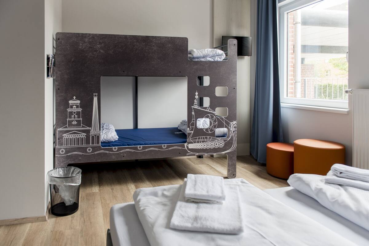 A bunk bed with a cityscape design, a window with curtains, and a small trash bin.