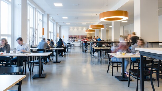 Una cafetería amplia con techos altos y ventanas grandes, que cuenta con luminarias modernas y diversas opciones de asientos.