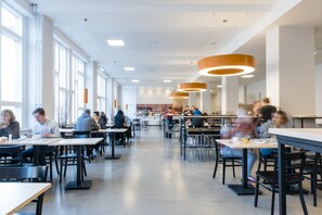 Una cafetería amplia con techos altos y ventanas grandes, que cuenta con luminarias modernas y diversas opciones de asientos.