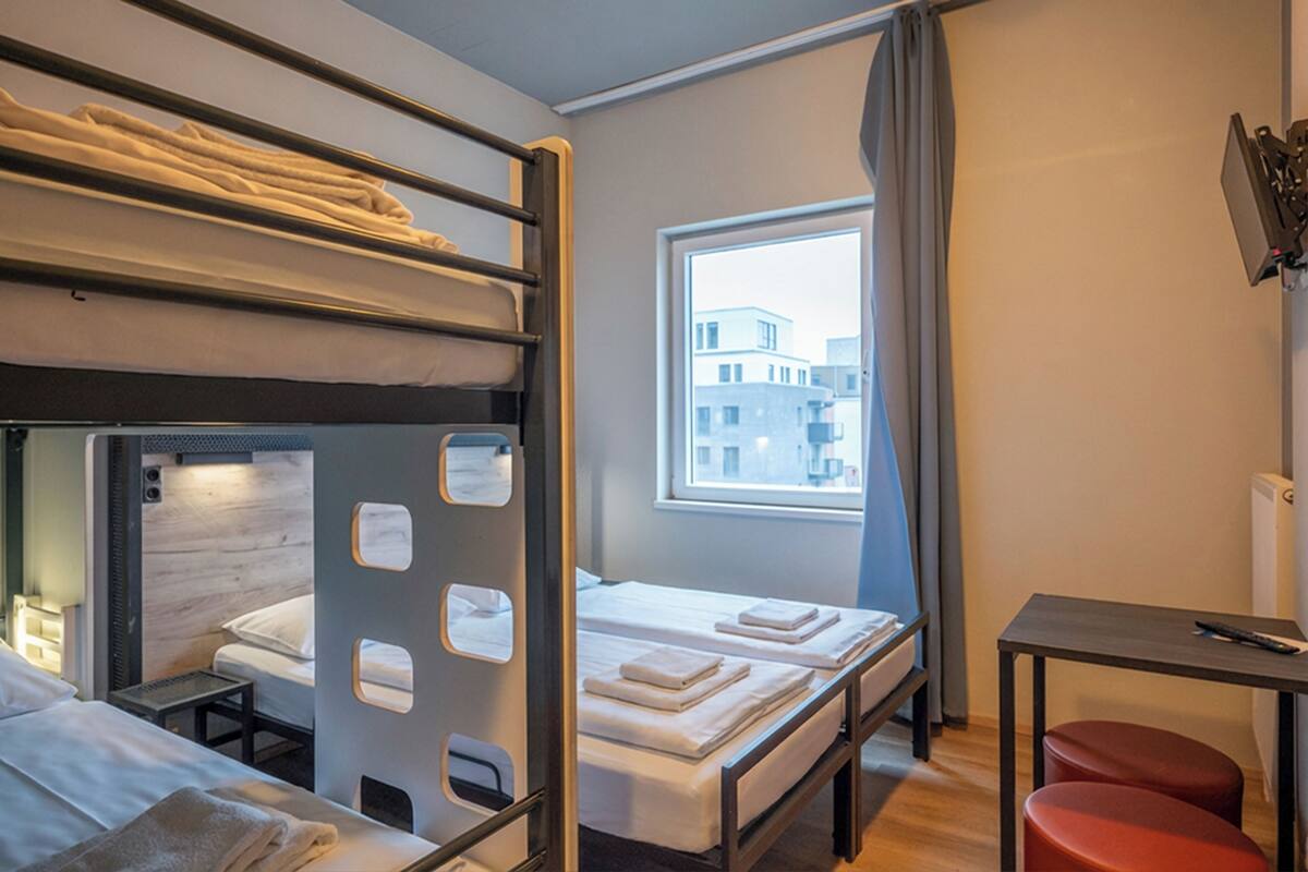A hotel room with bunk beds, a single bed, a small table, and a window with a view of buildings.