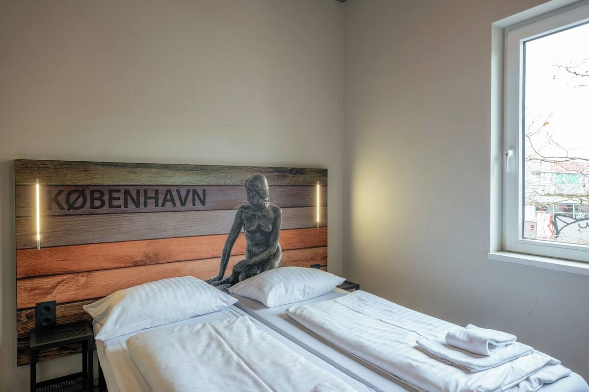A hotel room with a bed, a headboard with a sculpture, a nightstand, and a window with a view of the street.