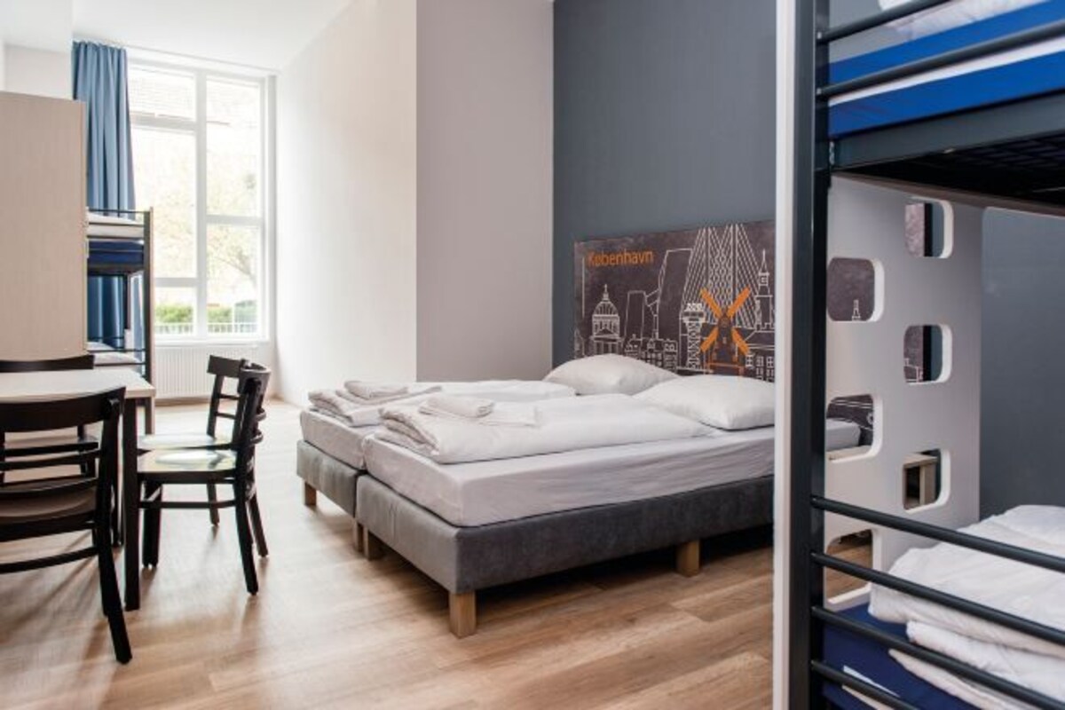 A modern hotel room with a bed, a desk, and a chair. There is a window with curtains, a poster on the wall, and a bunk bed in the corner.