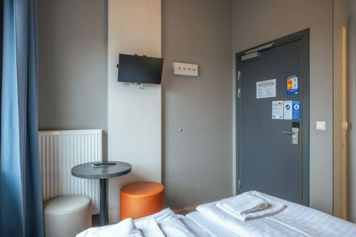 A hotel room with a bed, a small round table, a chair, a radiator, and a door with a bulletin board.