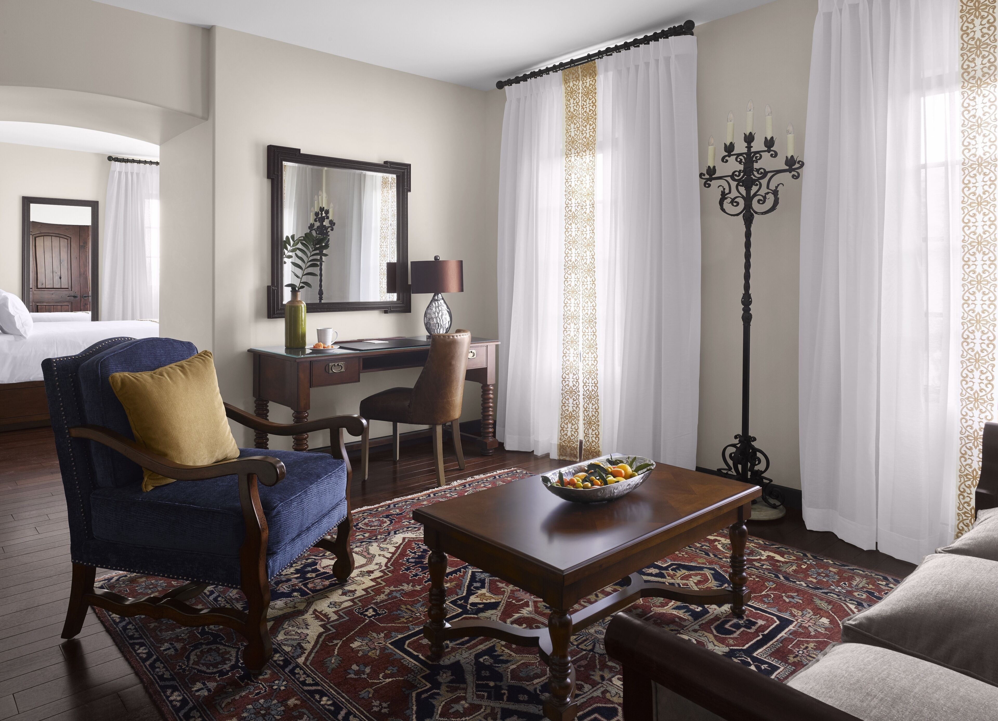 Habitación de hotel con cama, escritorio, silla, mesa con fruta, lámpara de pie y alfombra estampada.