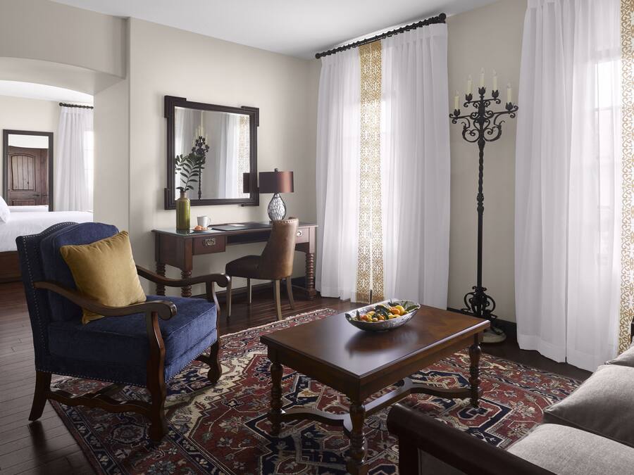 A hotel room with a bed, a desk, a chair, a table with fruit, a floor lamp, and a patterned rug.