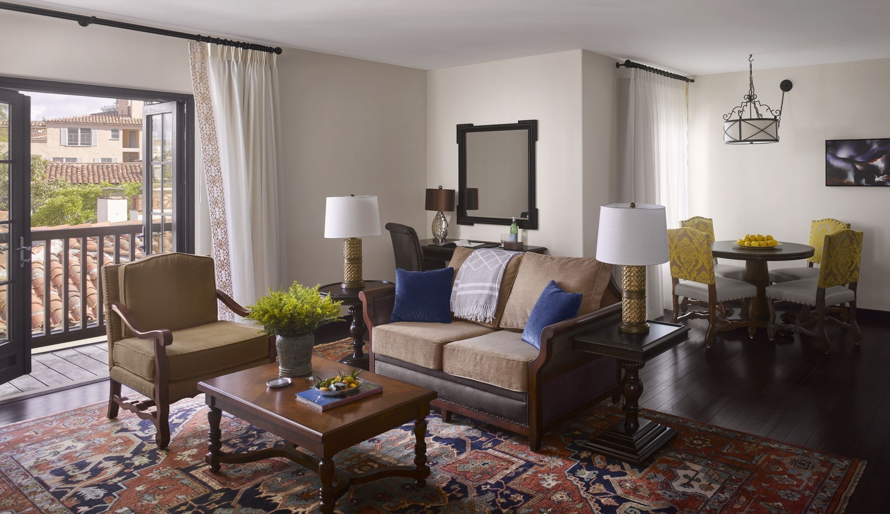 A living room with a sofa, armchairs, a coffee table, a dining table with chairs, a chandelier, and a balcony with a view of buildings.