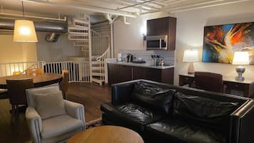 Un salon moderne avec un canapé en cuir noir, un fauteuil blanc, une table basse en bois et un coin repas avec une table et des chaises blanches. On y trouve également une cuisine avec des meubles sombres et un four à micro-ondes. Un escalier avec une rampe blanche mène à l’étage supérieur.