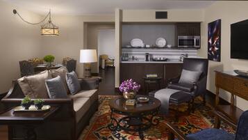 A living room with a sofa, armchairs, a coffee table, and a rug. There is a kitchen area with a microwave and cabinets.