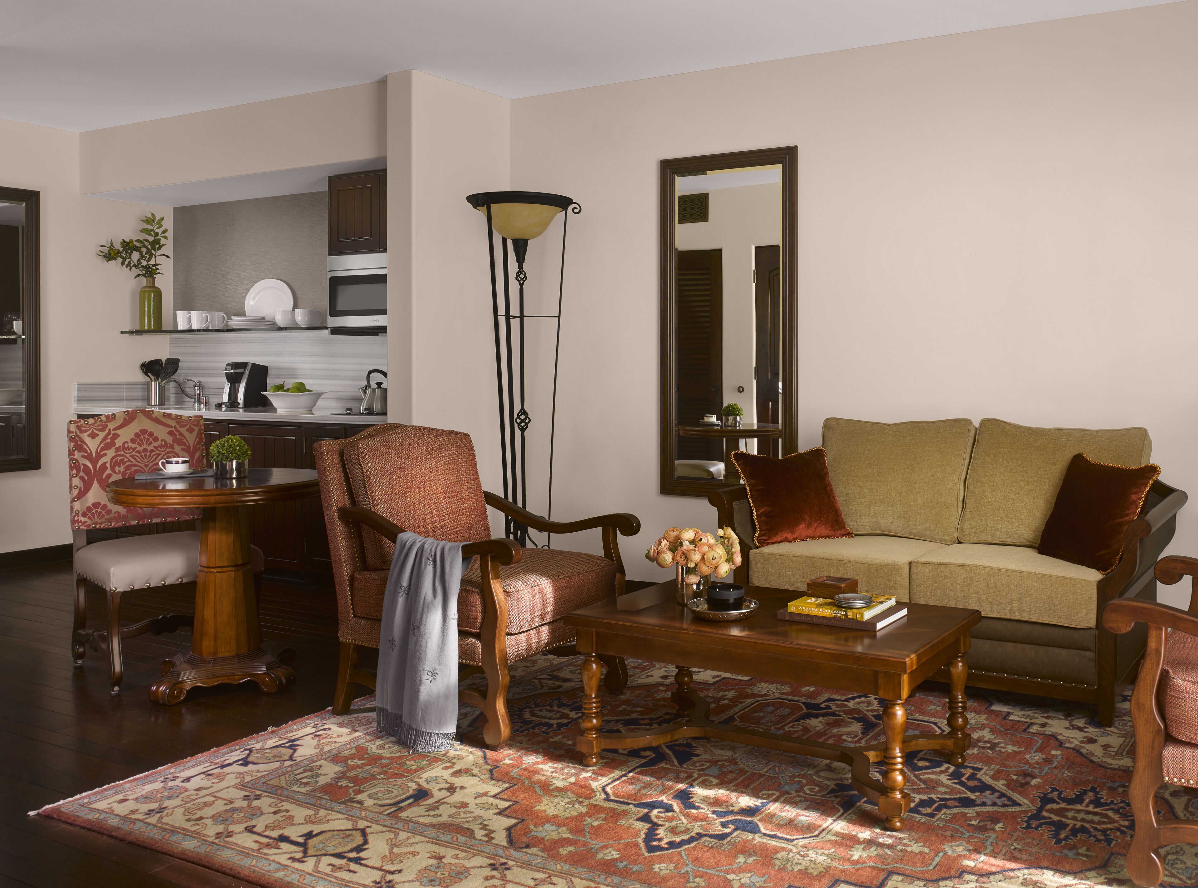 A living room with a sofa, armchair, coffee table, and a rug. There is a kitchen in the background with cabinets and a stove.