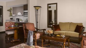 A living room with a sofa, armchair, coffee table, and a rug. There is a kitchen in the background with cabinets and a stove.