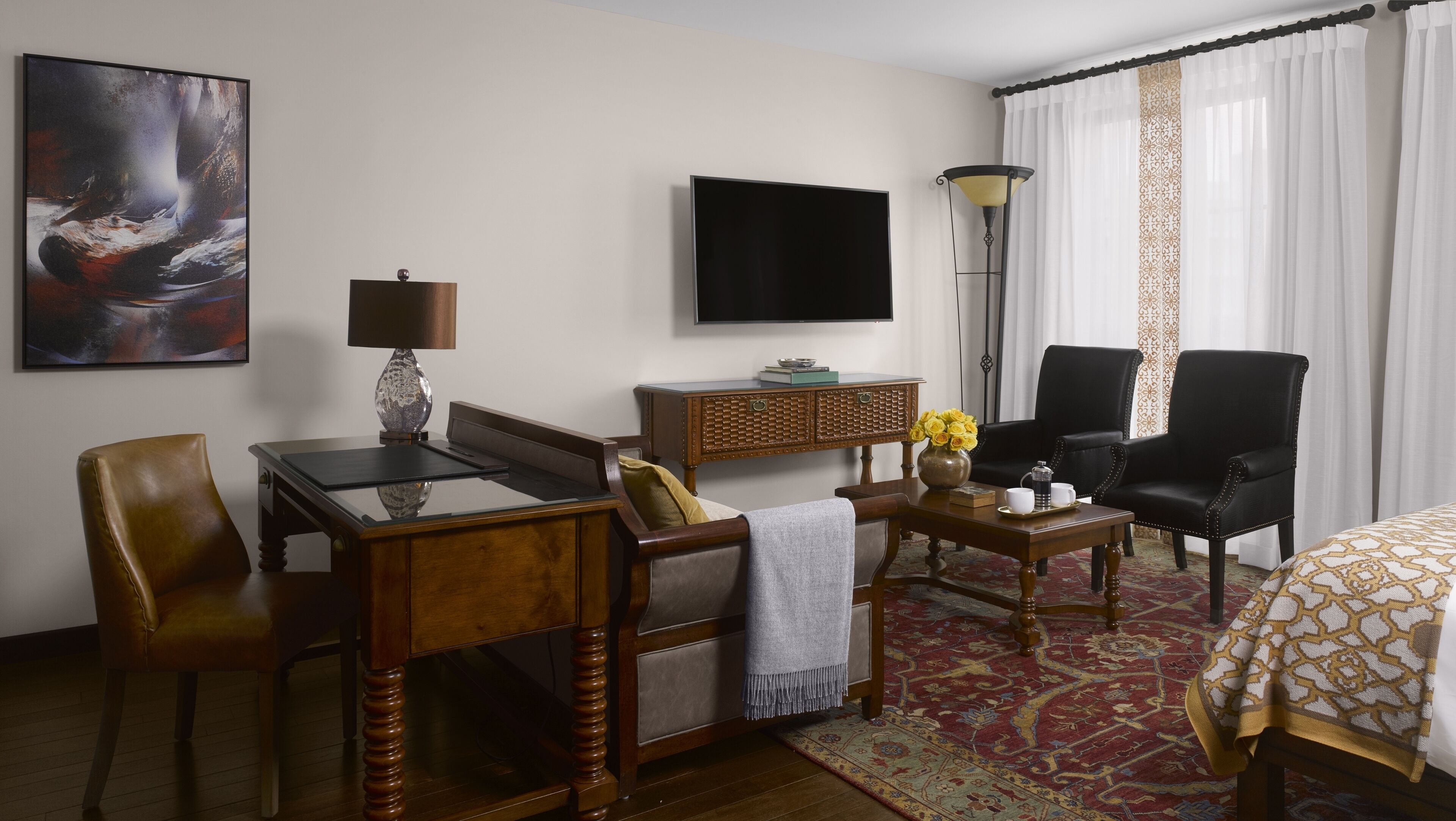 A hotel room with a wooden desk, a leather chair, a sofa, a flat-screen TV, a lamp, and a patterned rug.