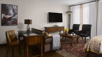 Habitación de hotel con escritorio de madera, una butaca de cuero, sofá, televisor de pantalla plana, lámpara y alfombra estampada.