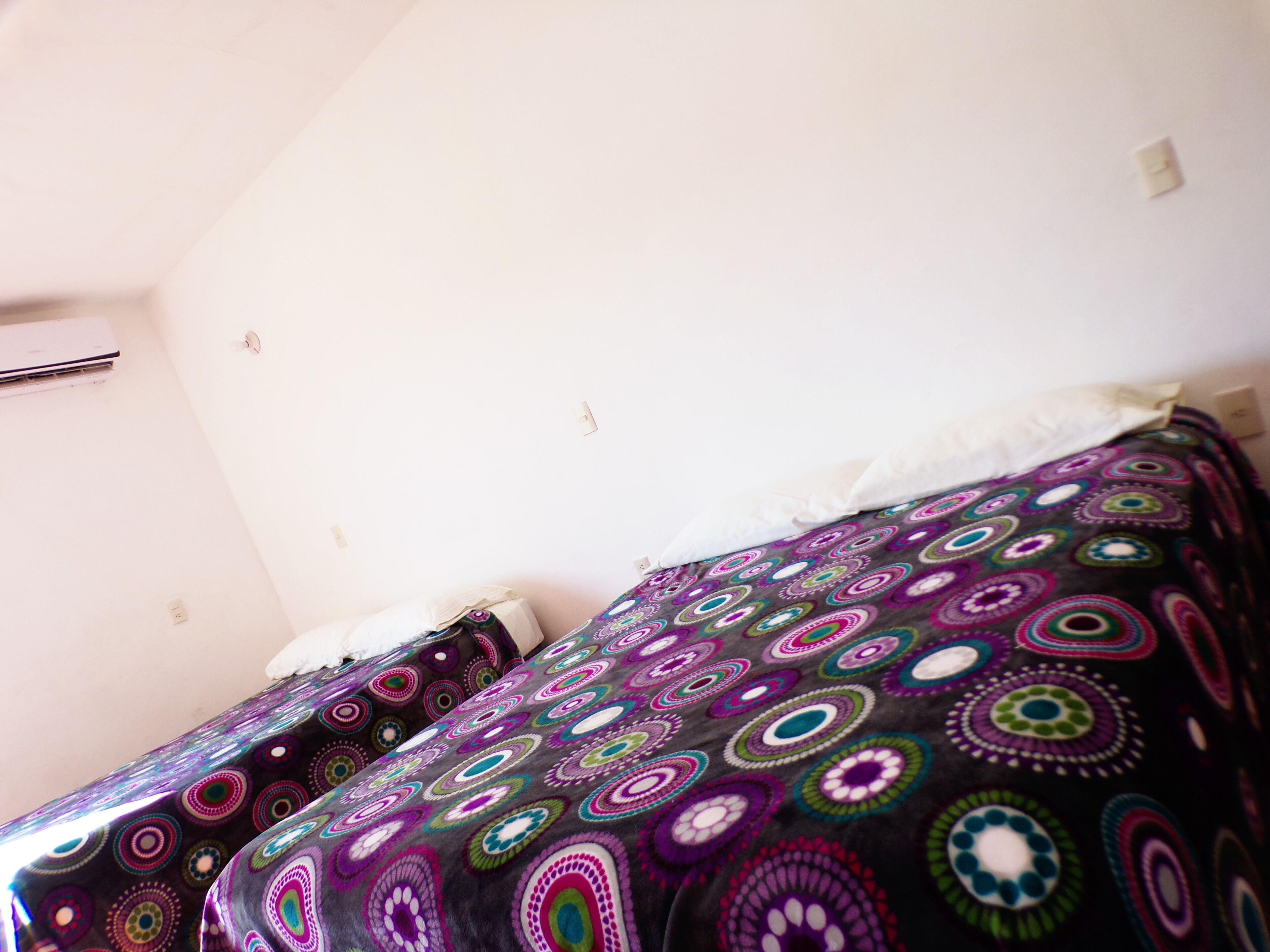 Une chambre à coucher avec un lit recouvert d’une couverture aux motifs colorés, un oreiller blanc et un mur blanc doté d’une prise de courant.