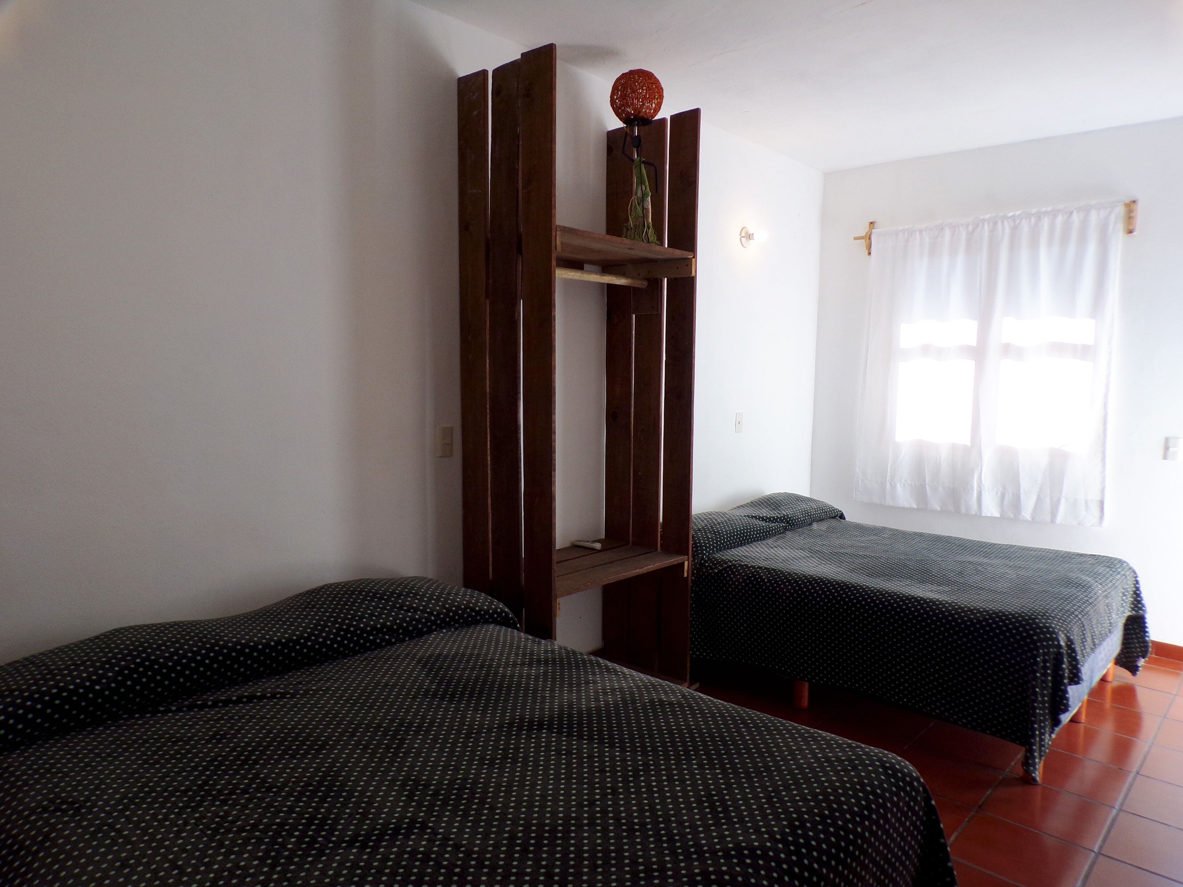 A room with two beds, a wooden shelf with a decorative item, and a window with curtains.