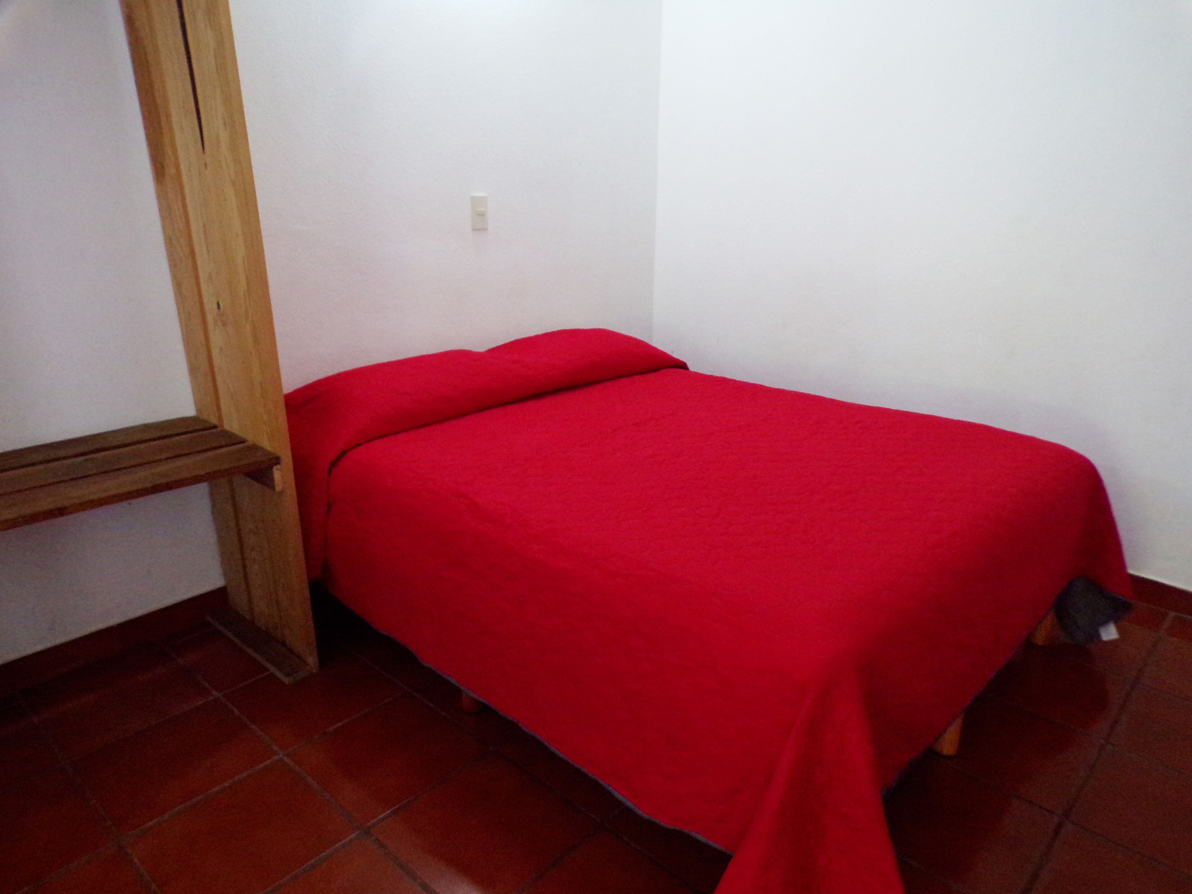 Ein kleines Zimmer mit einem Einzelbett, einer roten Tagesdecke, einem Holzkopfende und einem Fliesenboden.