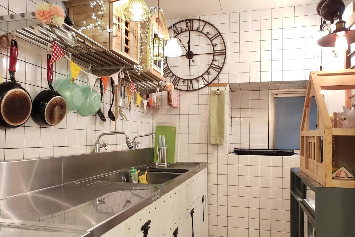 A kitchen with a large sink, hanging utensils, a clock, and a wooden shelf.