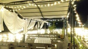 Un espace extérieur aménagé pour les repas, avec des guirlandes lumineuses, des rideaux blancs et des bancs en bois.