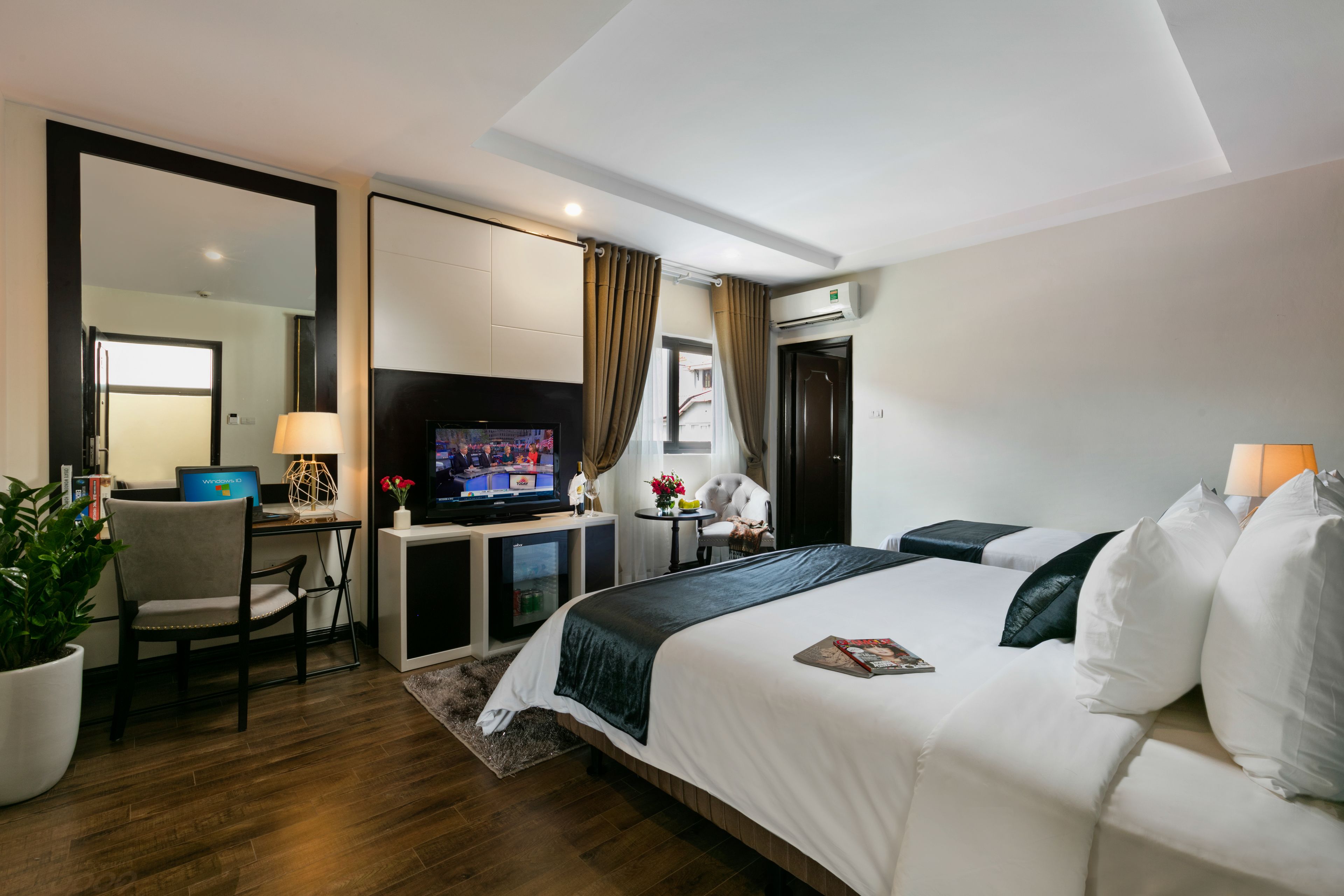 A modern hotel room with a large bed, a desk with a computer, a television, and a potted plant.