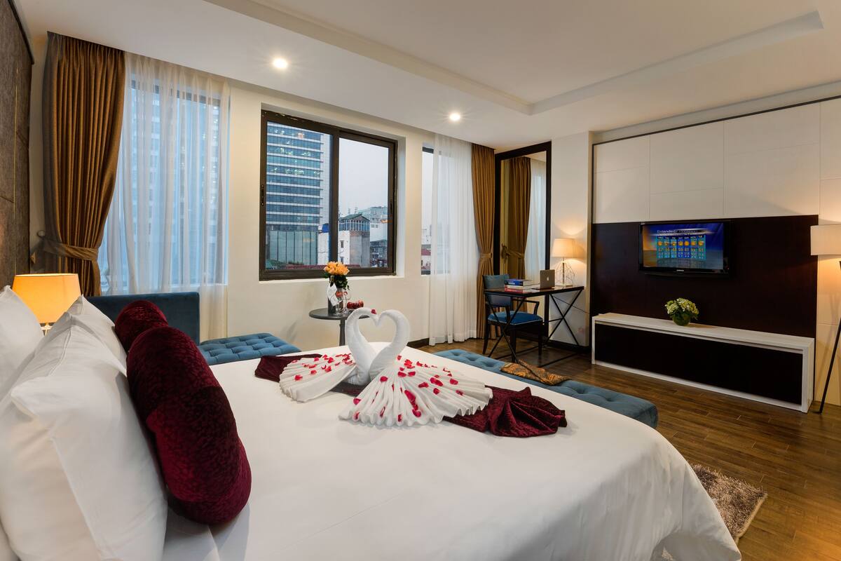 Habitación de hotel con una cama grande, televisión, una mesita con un florero y vistas a la ciudad desde la ventana.