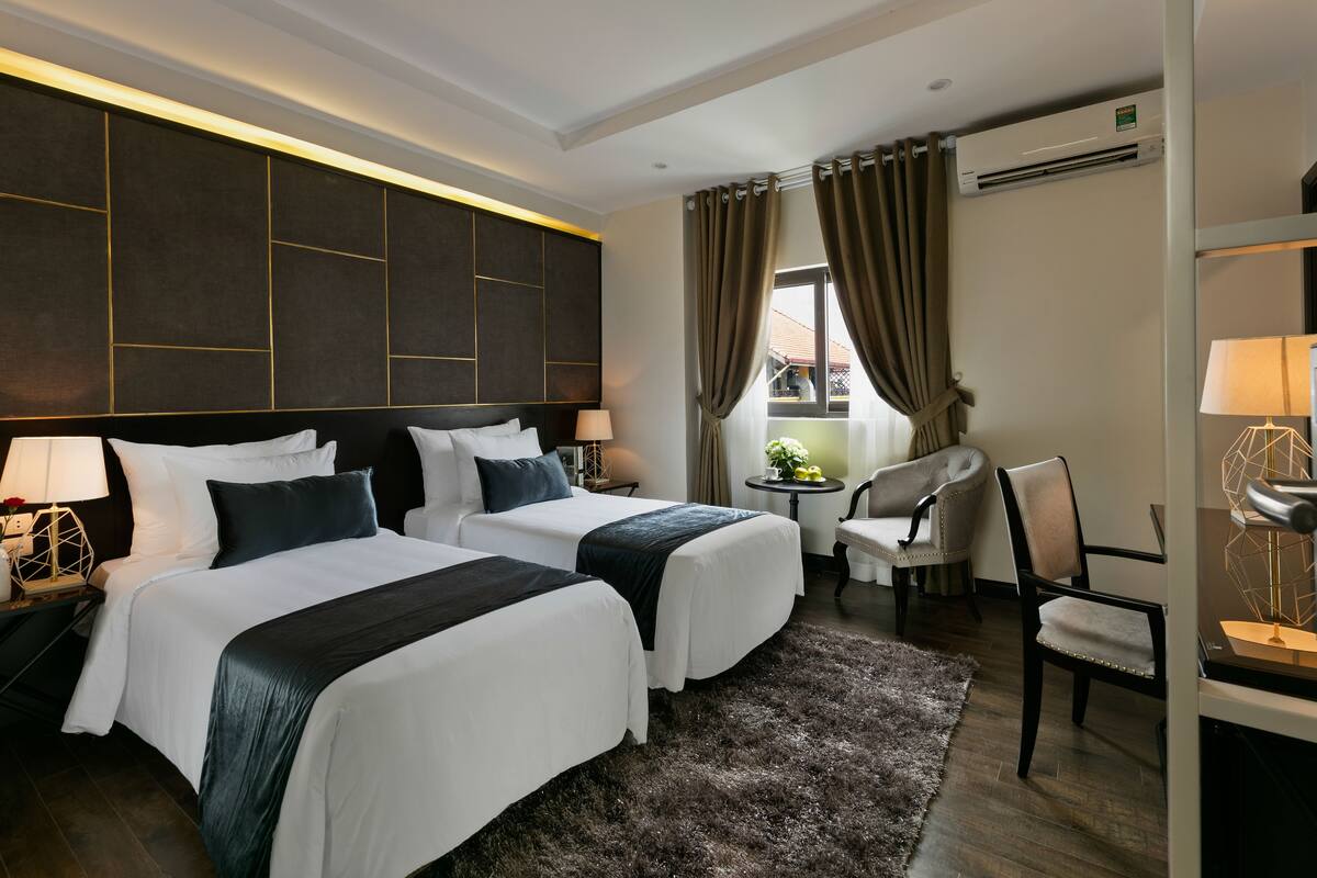 Habitación de hotel con dos camas, un escritorio, una silla, una mesita con lámpara y un aire acondicionado.