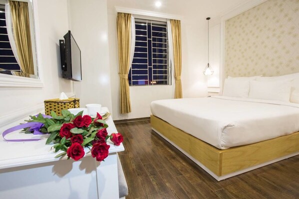 A hotel room with a bed, a bedside table with a bouquet of red roses, a television, and a window with curtains.