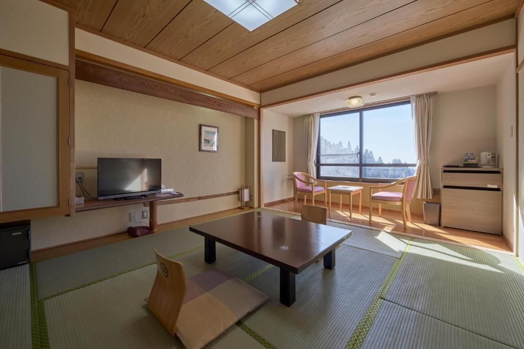 A hotel room with a wooden ceiling, a television, a dining table, and a view of a mountain through large windows.