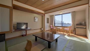 A hotel room with a wooden ceiling, a television, a dining table, and a view of a mountain through large windows.