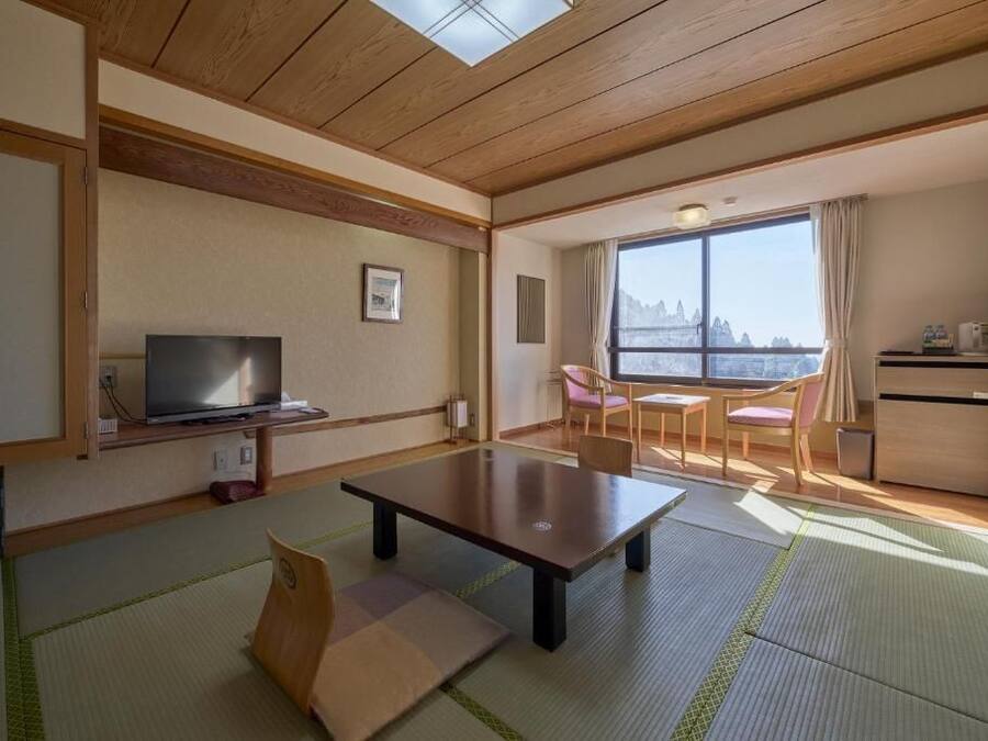 A hotel room with a wooden ceiling, a television, a dining table, and a view of a mountain through large windows.