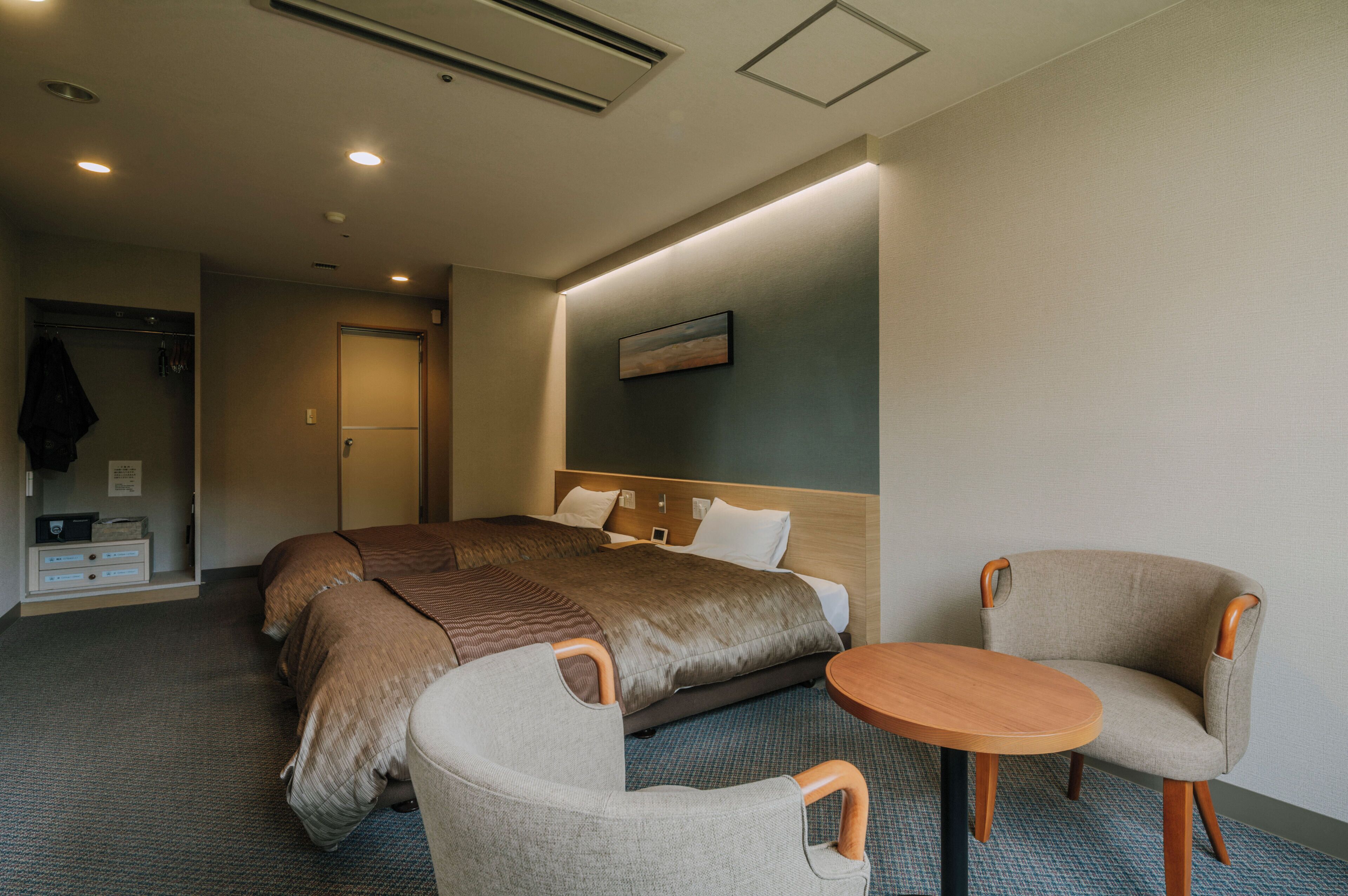Een hotelkamer met een groot bed, twee fauteuils, een klein houten tafeltje, een kapstok en een schilderij aan de muur.