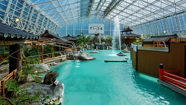 An indoor hot spring area with a glass ceiling, traditional wooden structures, and a central waterfall. - Kirishima Hotel (Kirishima)