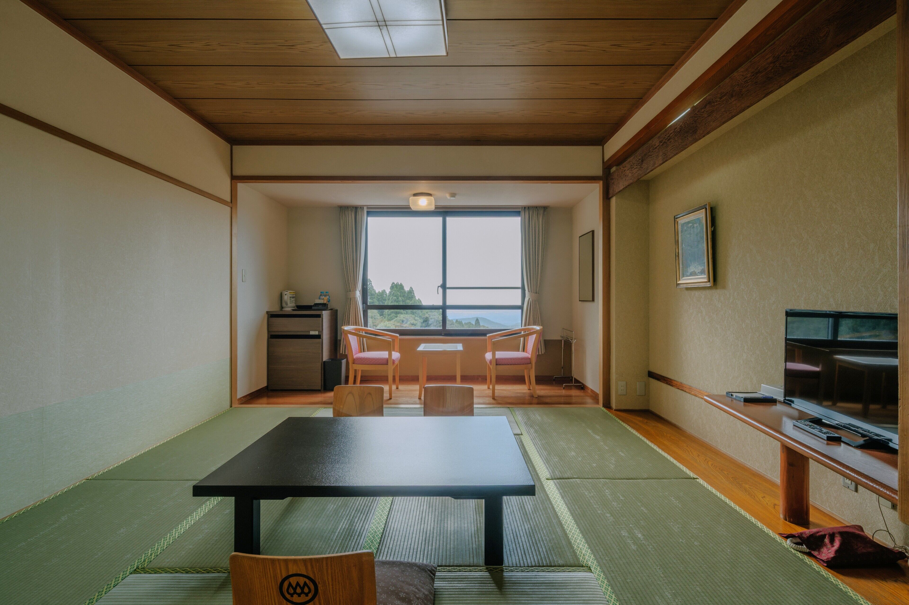 A traditional Japanese room with tatami flooring, a low table, two chairs, a TV, and a view through a large window.