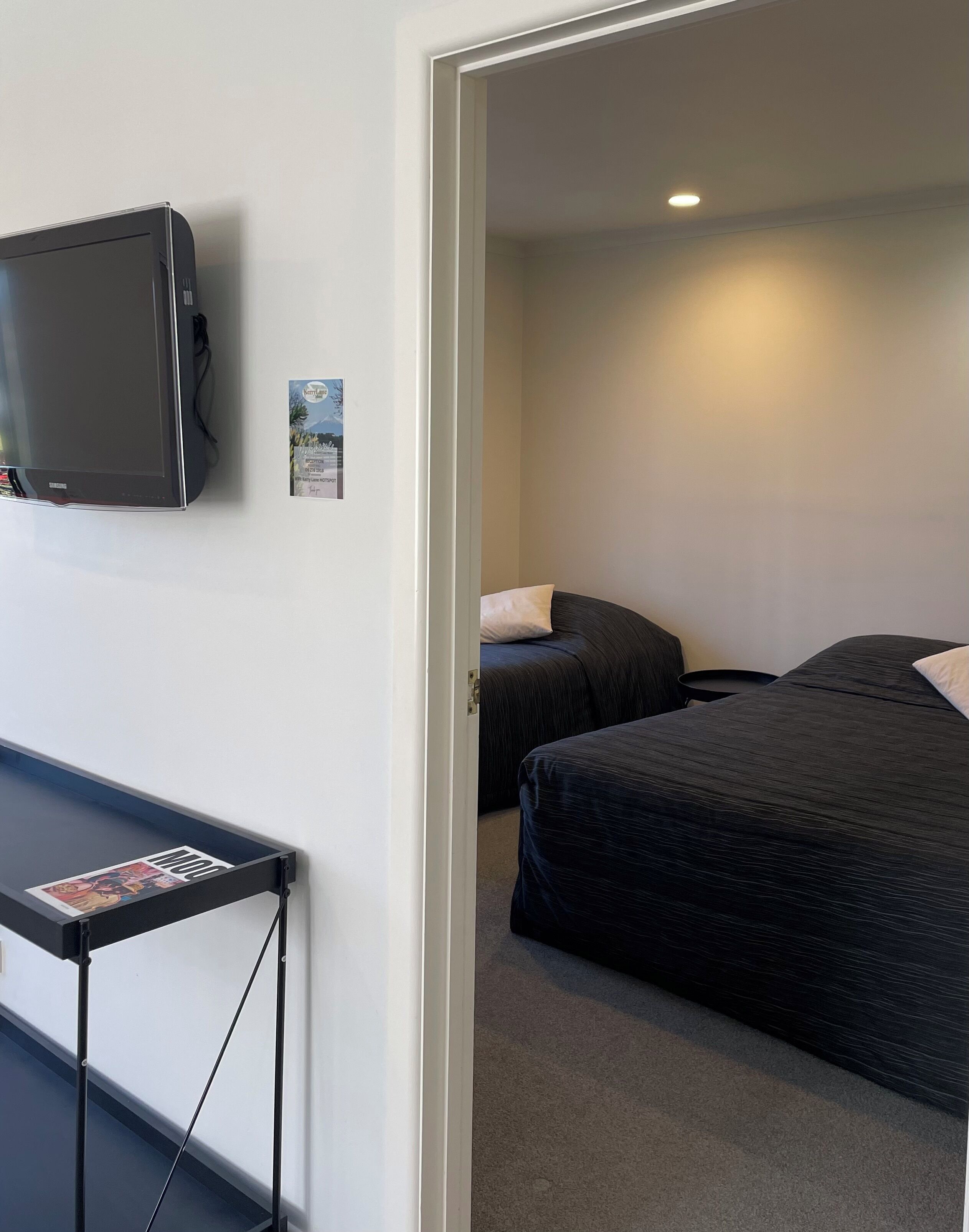A hotel room with a bed, a small table with a magazine, a television mounted on the wall, and a door leading to another room.