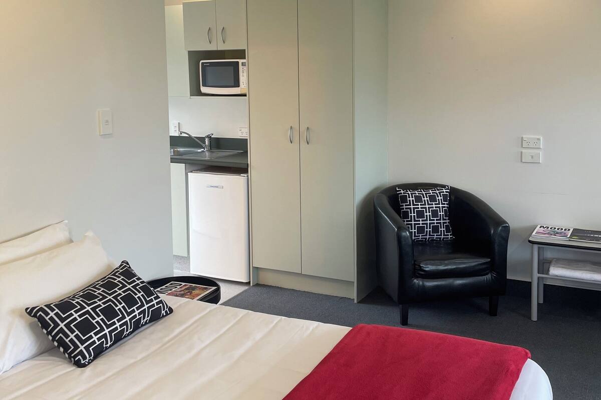 A hotel room with a bed, a desk, a chair, a kitchenette, and a microwave.
