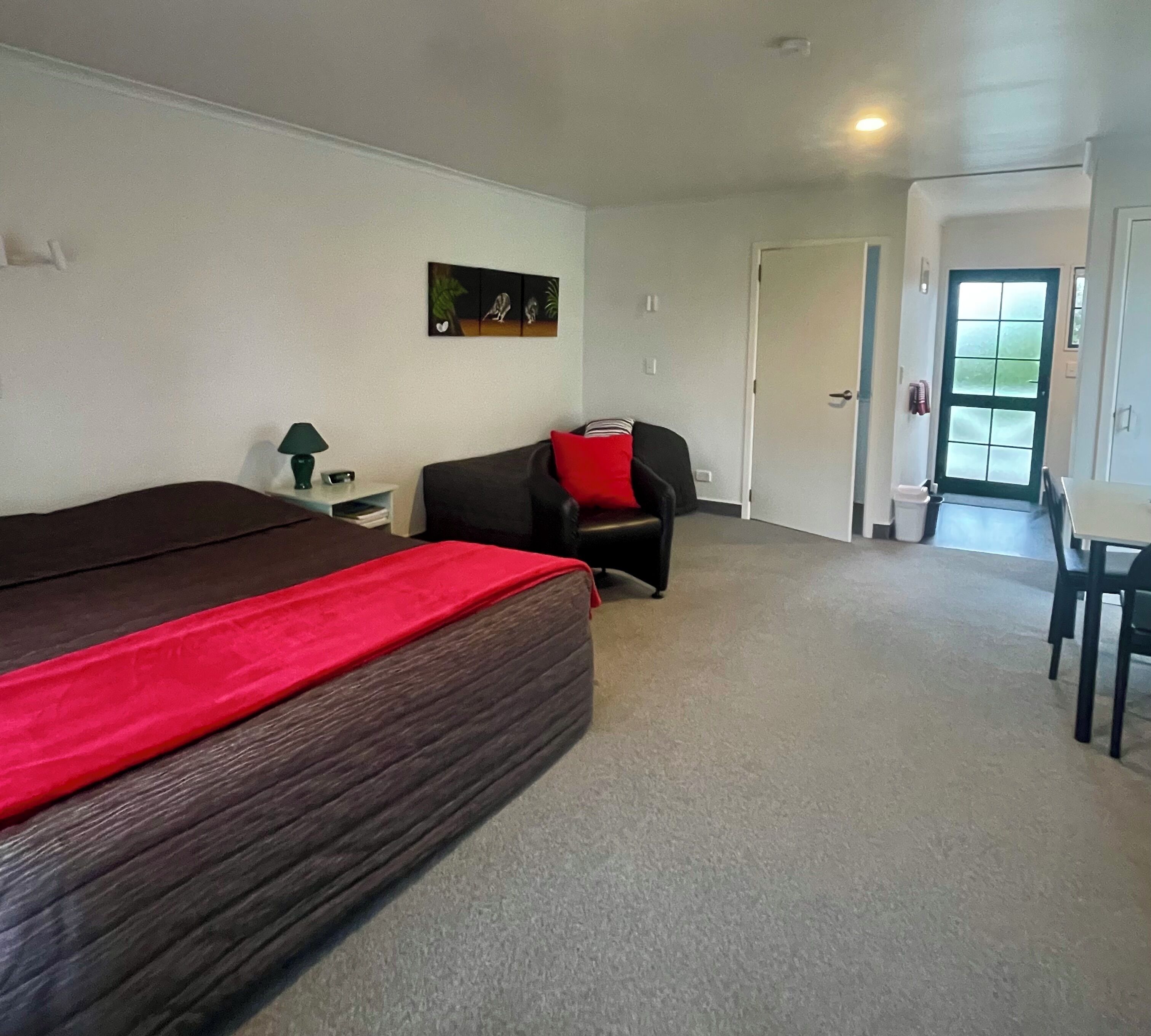 Una habitación con una cama grande, una silla con cojín rojo, una mesita y una puerta que da a otra estancia.
