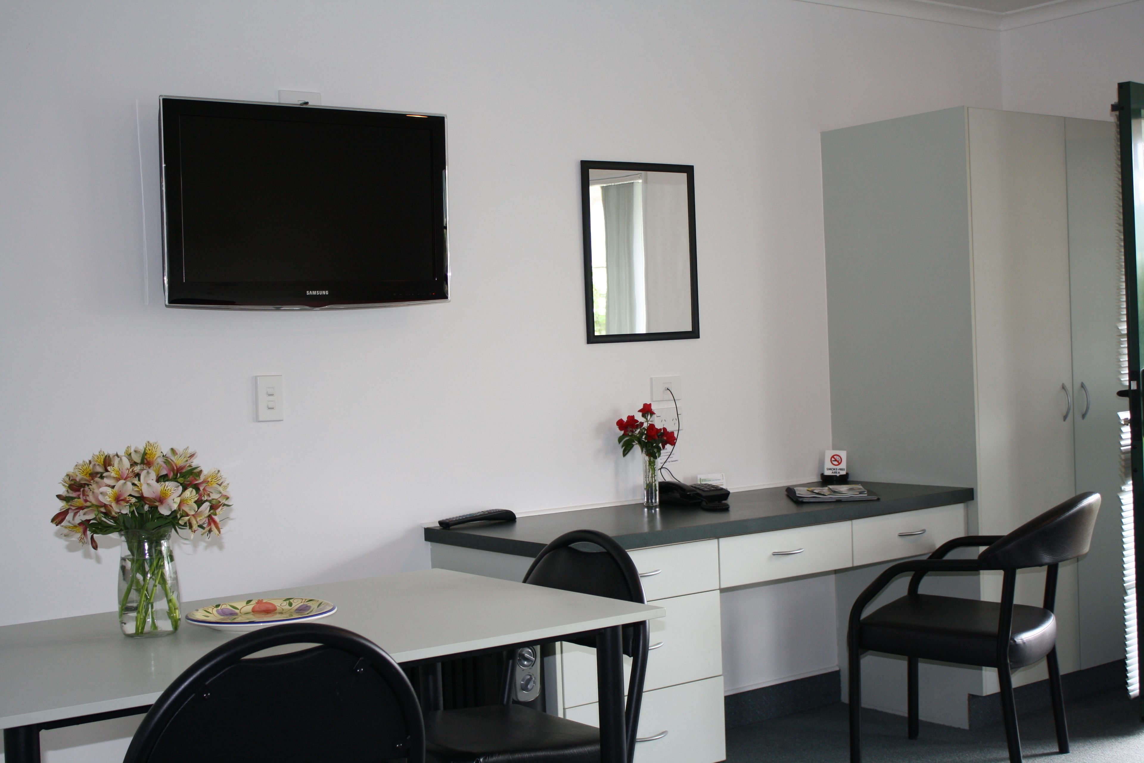 A modern room with a flat-screen TV, a mirror, a desk with chairs, and a vase with flowers.