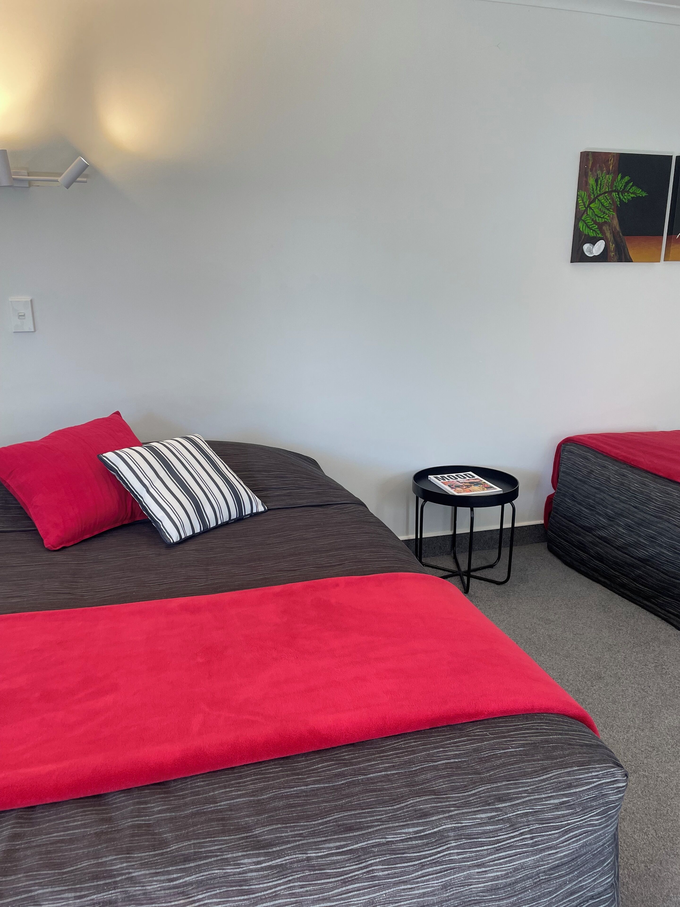 Una habitación con dos camas, una con una colcha roja y almohada a rayas, y otra con colcha roja. Hay una mesita redonda con una revista, y un cuadro en la pared.