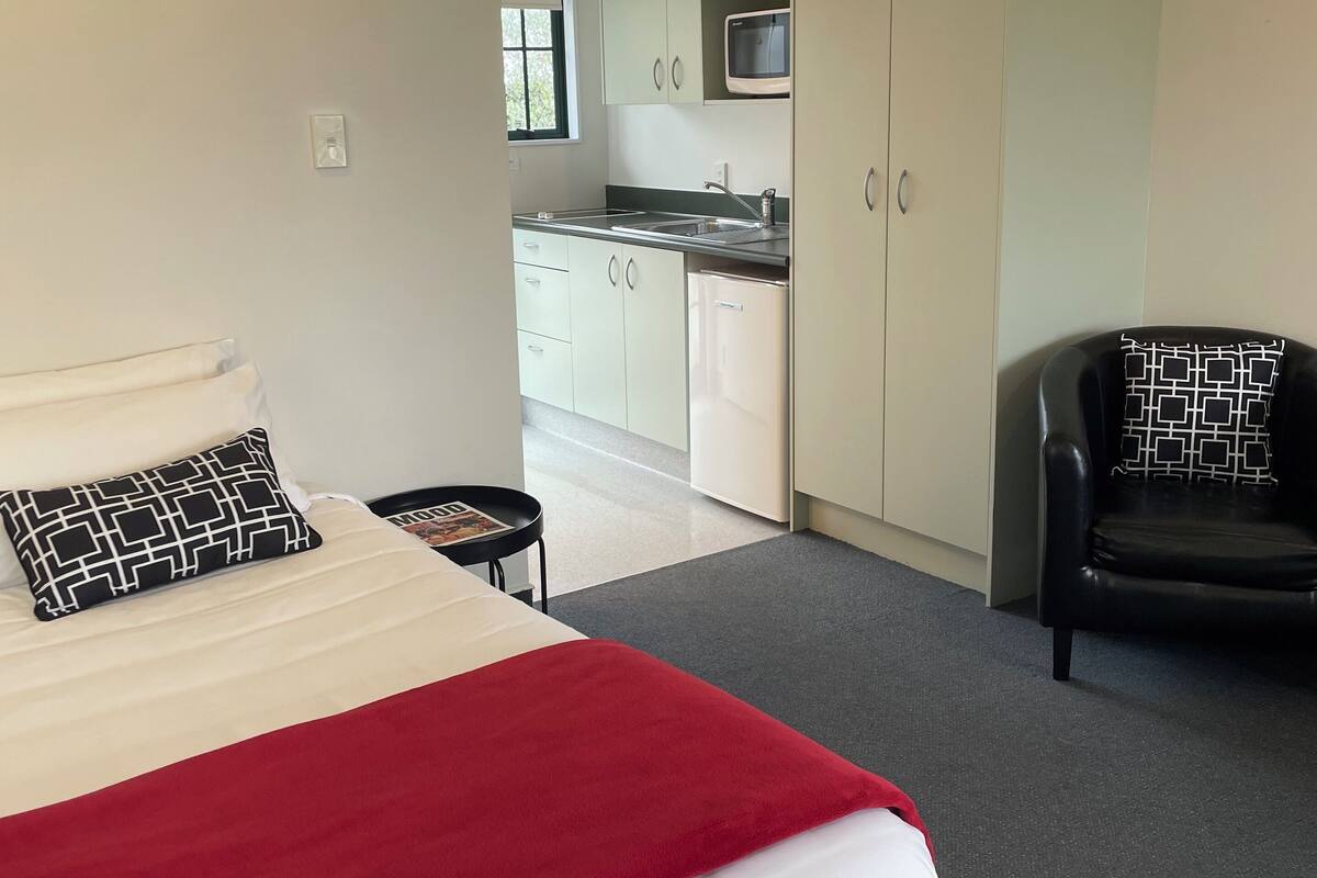 A bedroom with a bed, a chair, a small table, and a kitchenette in the background.