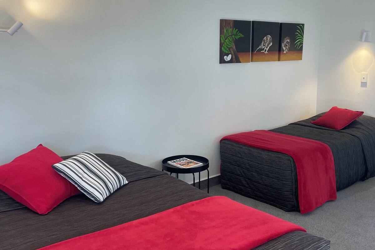 A modern hotel room with two beds, each with a red blanket and a striped pillow. There is a small round table with a magazine, and a wall-mounted light fixture.