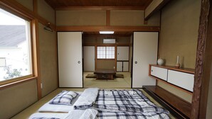 Une pièce de style japonais traditionnel, avec un sol recouvert de tatamis, un futon servant de lit, des portes coulissantes et une vue sur une autre pièce de style japonais à travers la fenêtre.