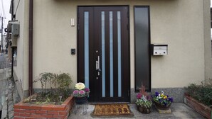 Une entrée d’immeuble avec une porte sombre, un paillasson et des plantes en pot.