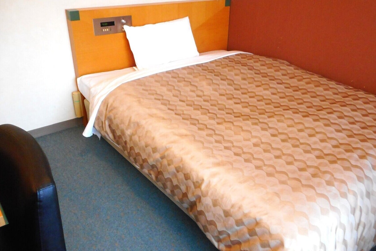 Uma cama de solteiro com um cobre-leito estampado, um travesseiro branco e um relógio digital na cabeceira.