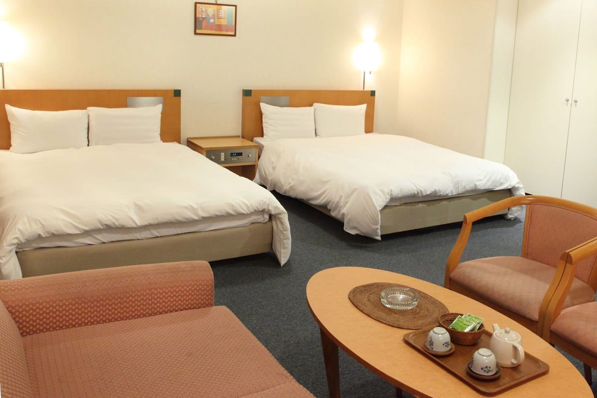 Quarto de hotel com duas camas, uma mesinha e um conjunto de chá, além de uma cadeira.