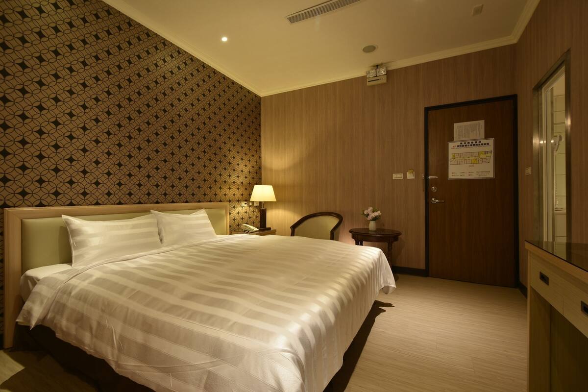 Una camera d'albergo con un letto grande, un comodino con una lampada, una sedia, una porta in legno e uno specchio.