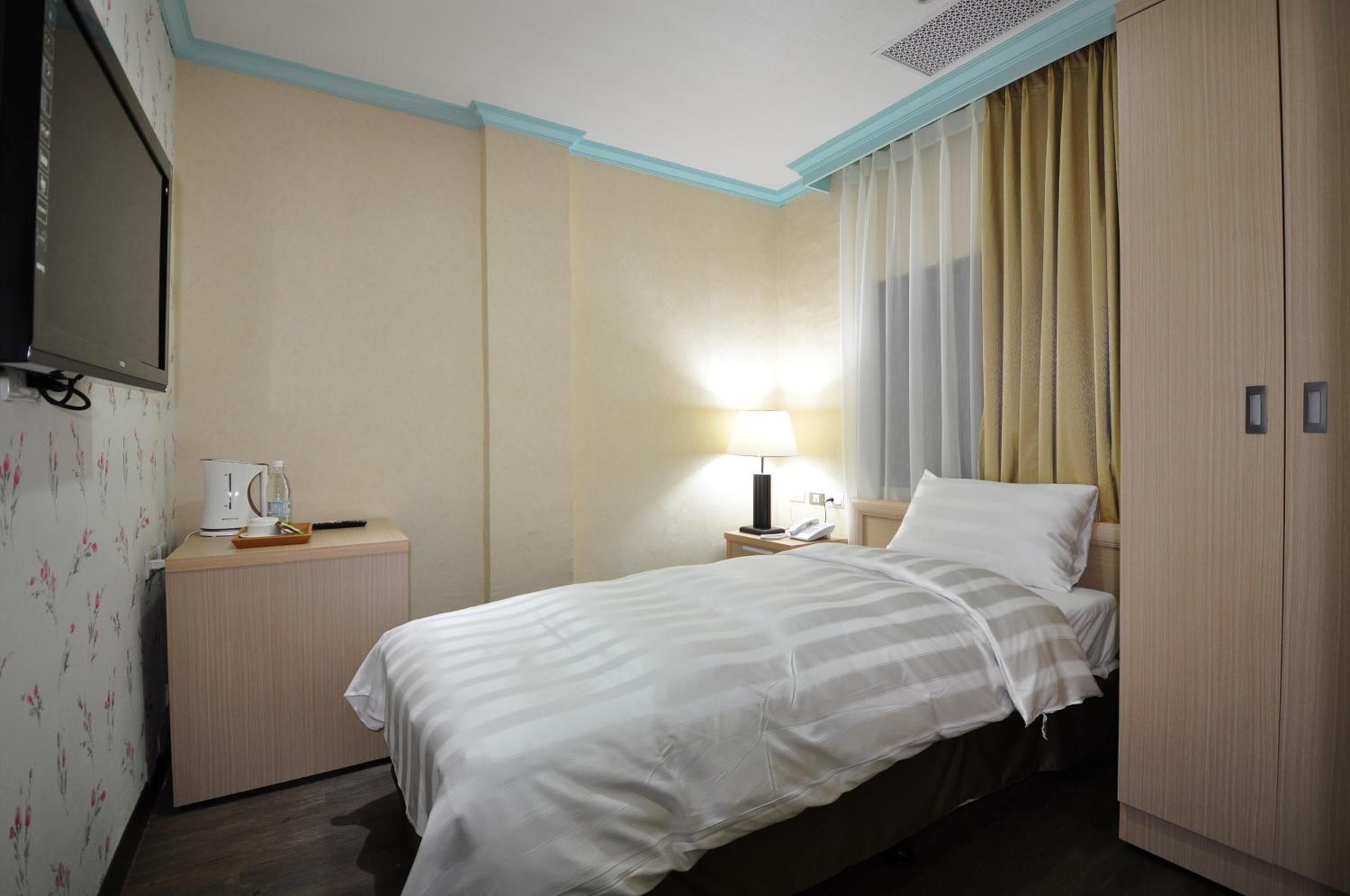 Quarto de hotel com cama, mesa de cabeceira, luminária, televisão e guarda-roupa.