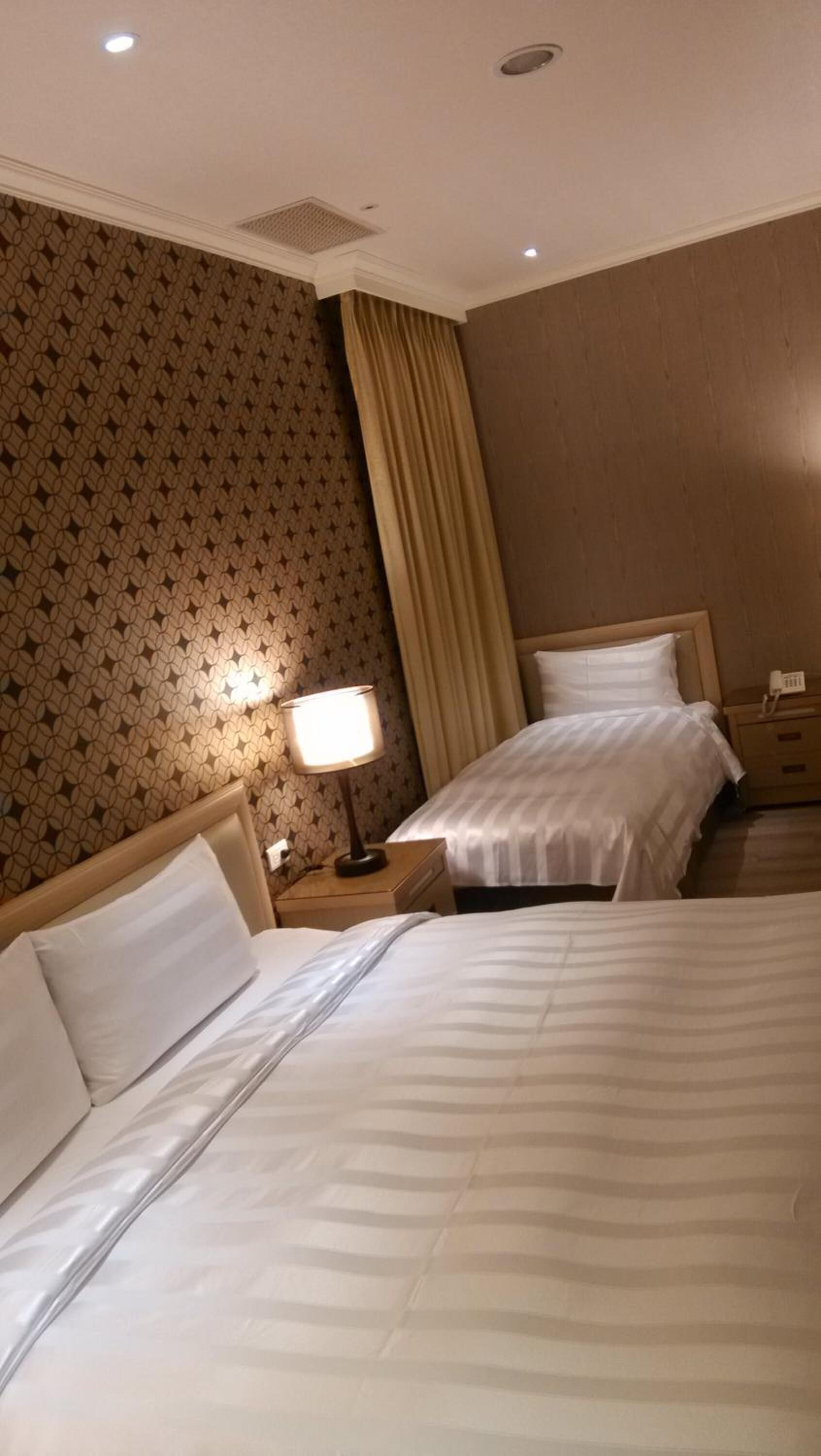 Habitación de hotel con dos camas, una mesita de noche con lámpara y un papel tapiz estampado.